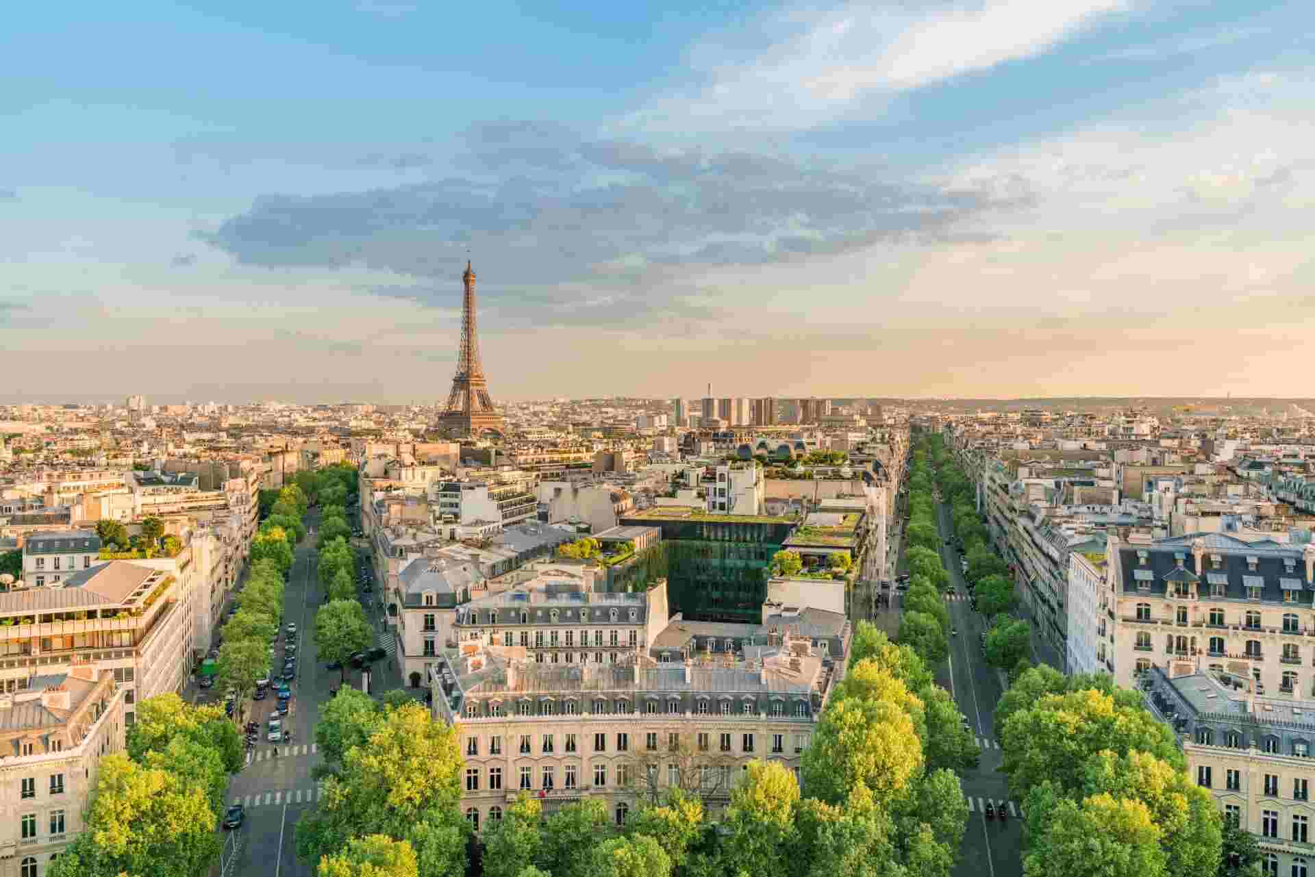 Property in France Wide aerial view of Paris with the Eiffel Tower centered in the distance, surrounded by historic buildings and green tree-lined streets under a partly cloudy evening sky.