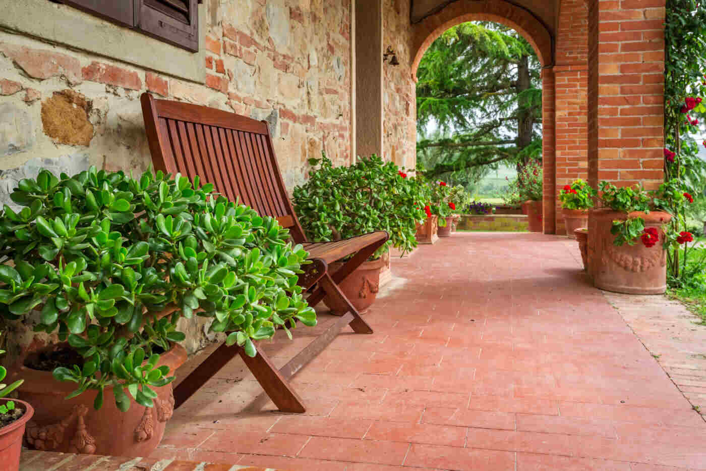 Holiday home owner in France? Choose a reliable property tax filing assistance! A scenic outdoor terrace with terracotta tiles, stone walls, and a wooden chair surrounded by potted green plants. A brick archway leads to a garden with large evergreen trees.