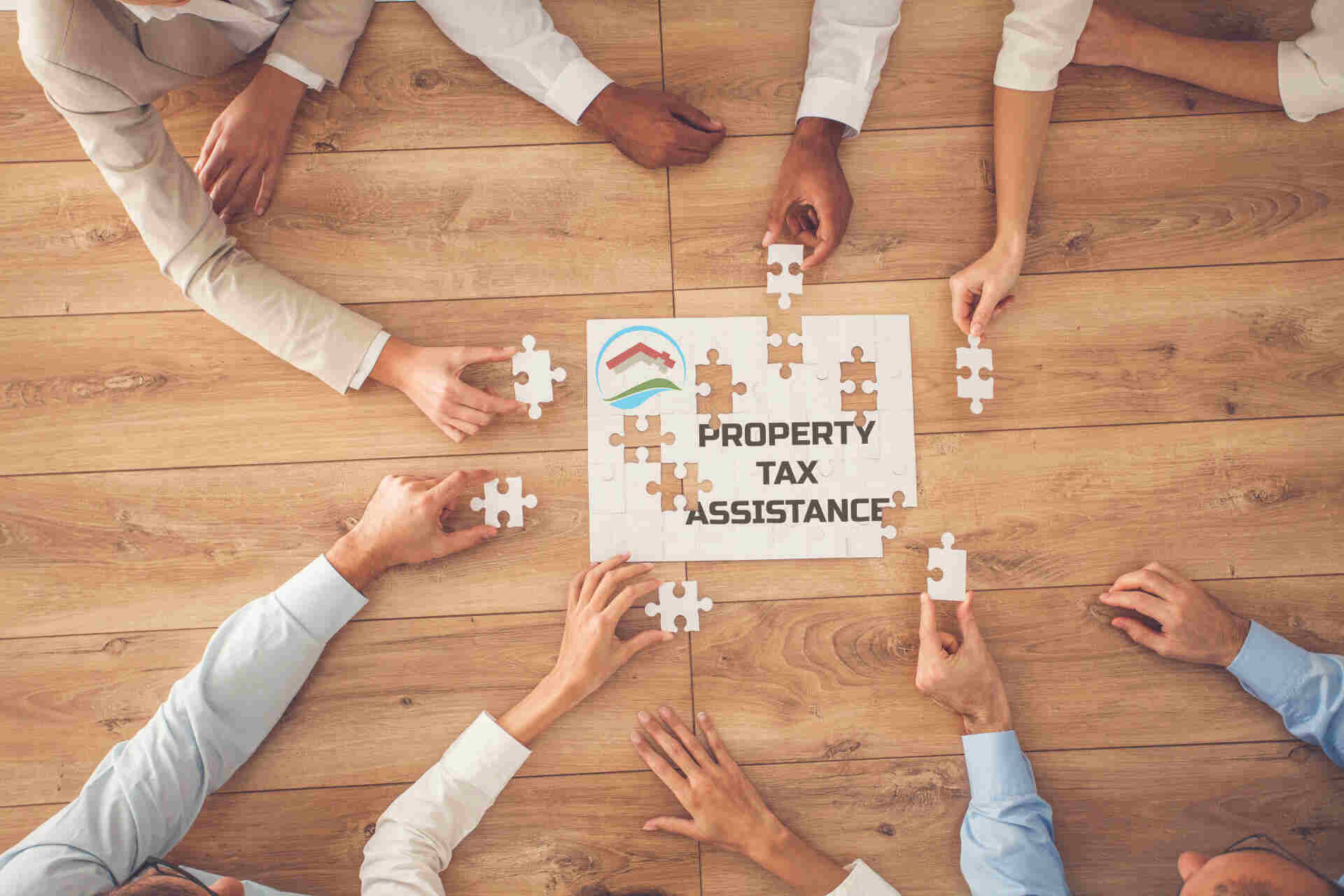 Property tax return assistance A group of professionals collaborate around a table, assembling puzzle pieces around a central sign that reads “Property Tax Assistance.”