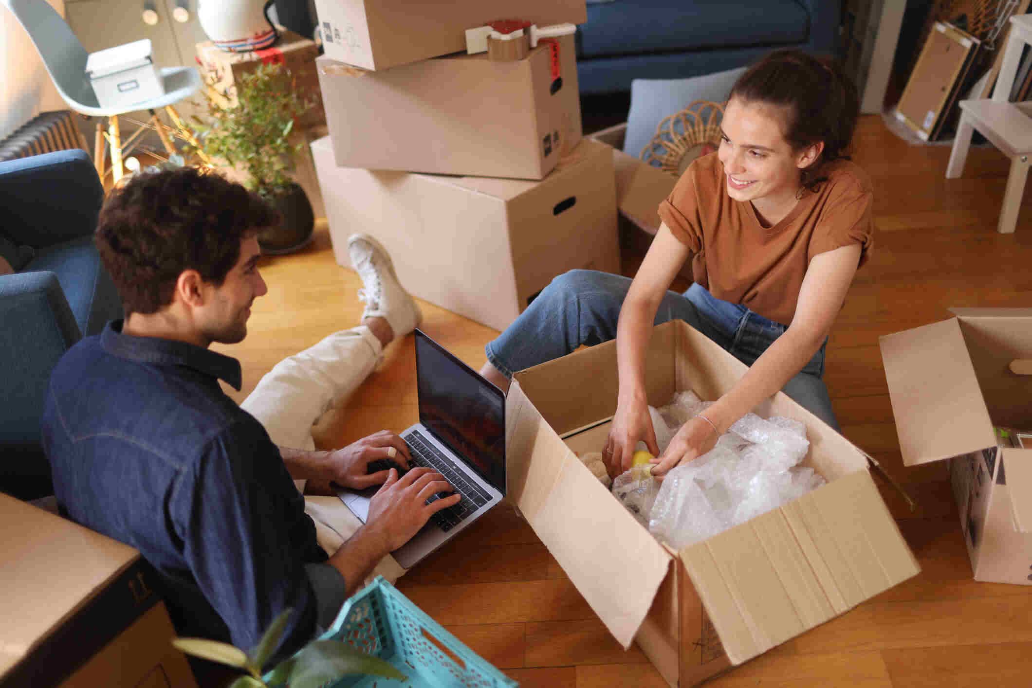 A smiling young couple carrying boxes and a plant into their new modern French apartment, highlighting the excitement of a new home purchase.